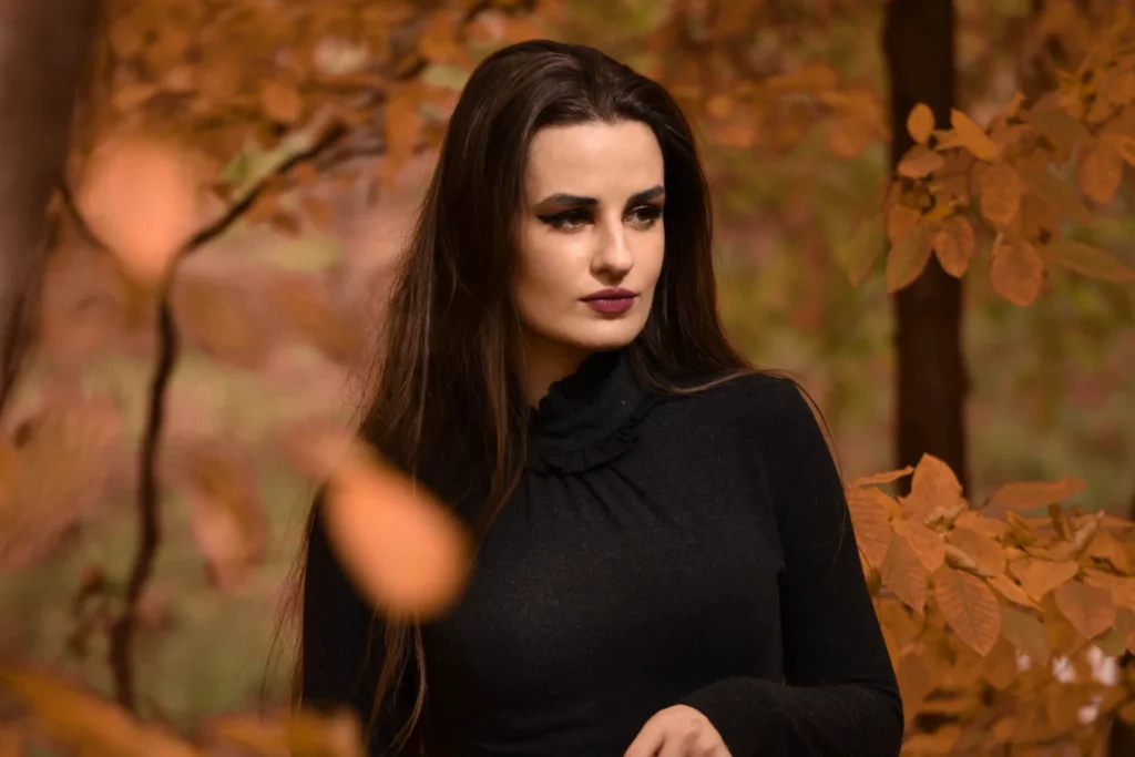 Młoda kobieta w czarnej sukience pozuje w jesiennym lesie, otoczona drzewami i liśćmi.