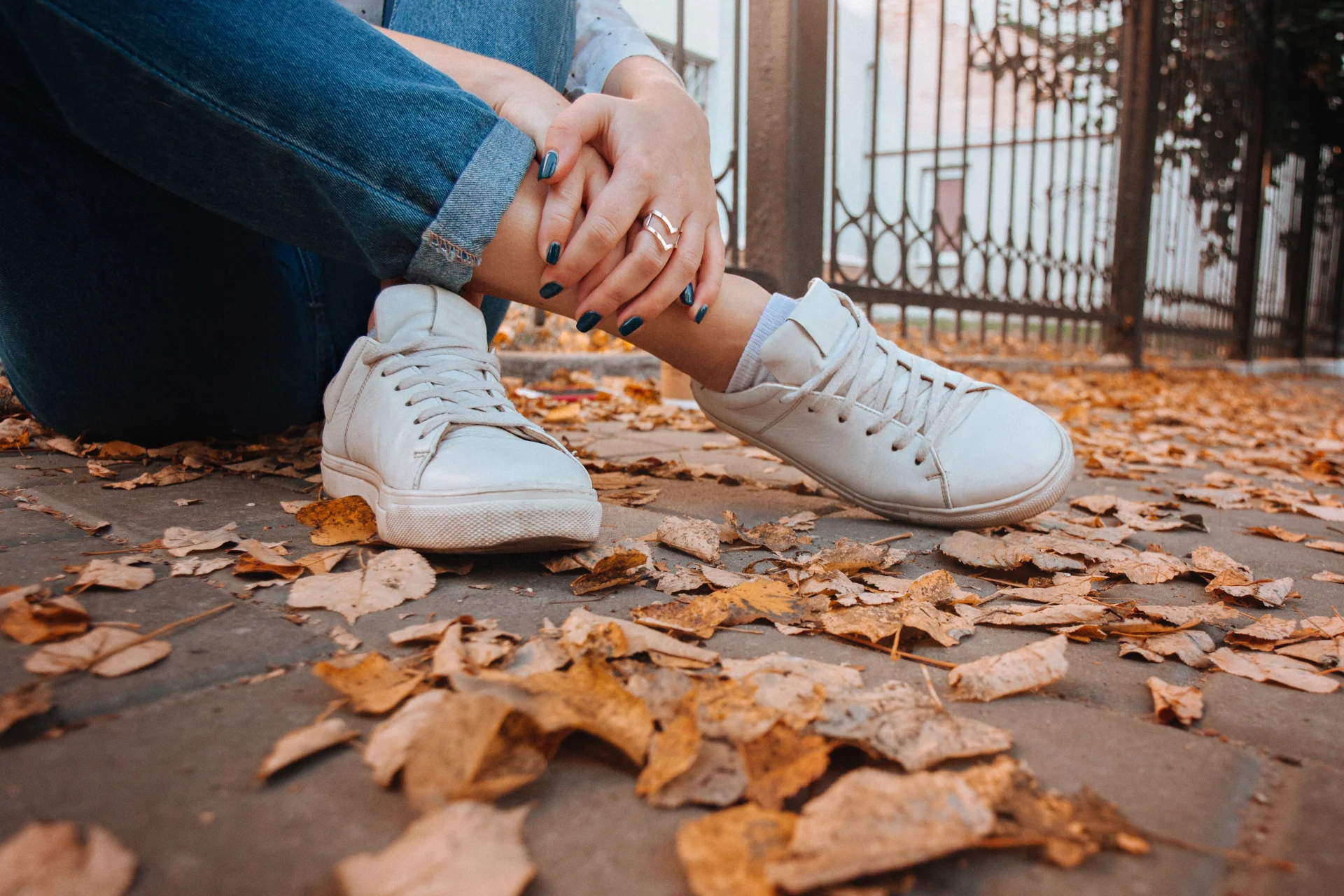 Zbliżenie na kobietę w białych sneakersach siedzącą na chodniku pośród jesiennych liści.